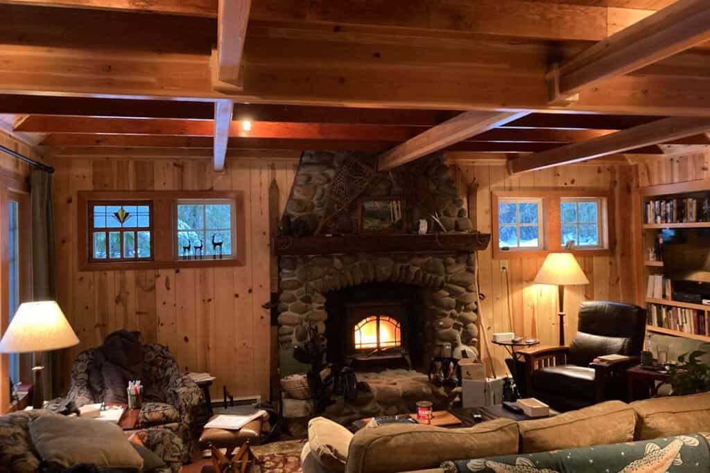 A cozy cabin living room with a fire burning in the wood stove. The living room has pine paneling and an exposed wood beam ceiling.