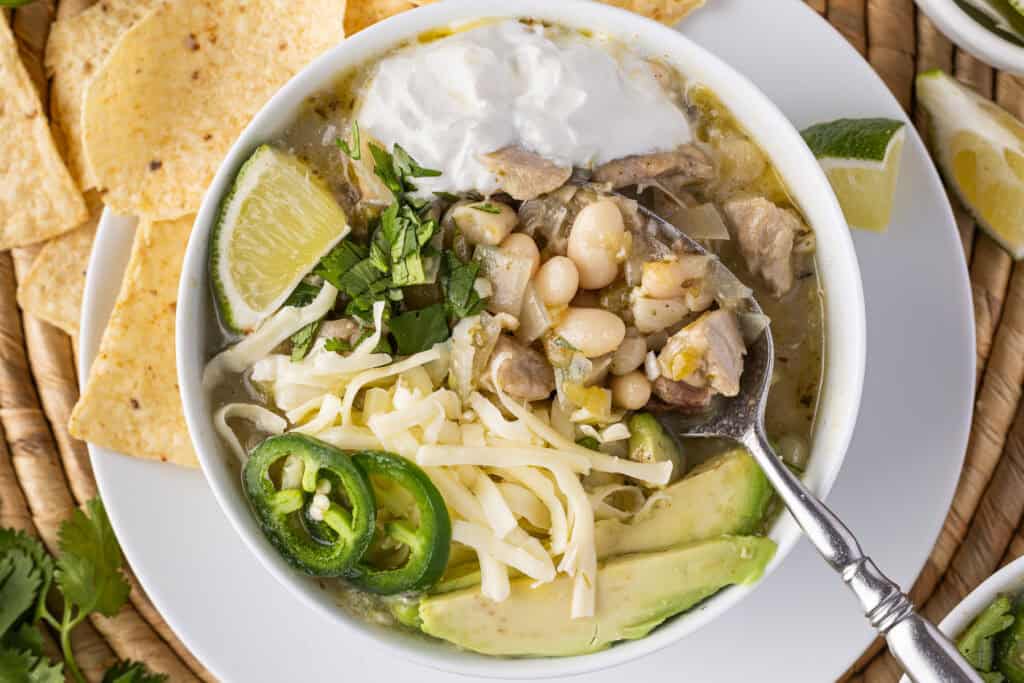 Bowl of White bean chicken chili with a spoon in it. The chili is garnished with sour cream, lime, Monterey Jack cheese, avocado cilantro and jalape&ntilde;o. There are tortilla chips on the side.