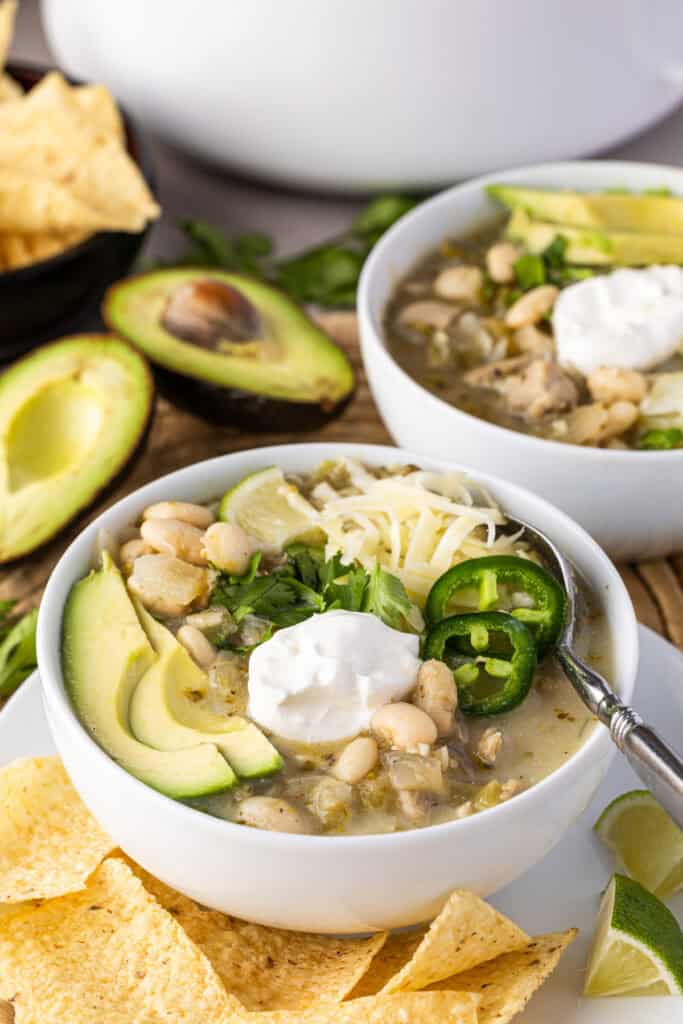 Bowls full of White Bean Chicken Chili with garnishes on top. Avocado, sour cream, cilantro, jalape&ntilde;os and cheese. There are avocados and chips on the table.