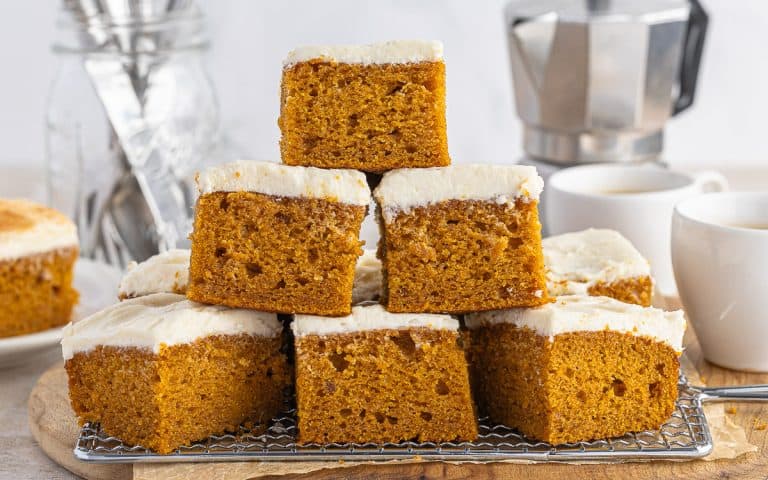Stacked pumpkin bars with cream cheese frosting show the moist, fluffy interior. Coffee pot in the background and a cup of coffee on the side.